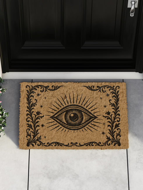 A 24x16 inch rectangular outdoor doormat made of eco-friendly coconut coir with a black vinyl backing. The mat features a central evil eye design radiating with detailed rays and surrounded by swirling ornamental scrollwork and small stars. Designed for witchy home entryways, it offers protection and style for porches, patios, and thresholds.